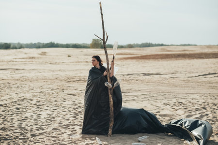 Environmental protection. Plastic reduction. Sustainable Earth. Confident mad woman wearing black trash bag standing embracing dry tree with used empty bottles in sandy desert with cloud skyline.の写真素材