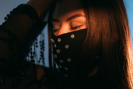 Pandemic fashion. Glamorous elegance. New normal style. Asian woman in black dress and decorated crystal beads face mask closing eyes posing on blue light shadow.の写真素材