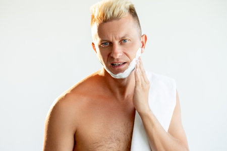 Male grooming. Barber shop. Face care treatment. Portrait of brutal confident shirtless blond man applying white shaving foam on chin isolated on light empty space background.の写真素材
