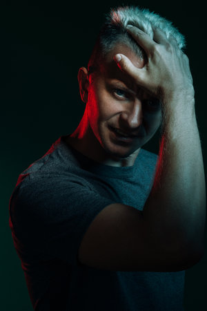 Macho lifestyle. Neon light portrait. Male confidence. Strong athletic man model in t-shirt looking at camera posing showing biceps combing hair with hand in red glow isolated on dark teal background.の写真素材