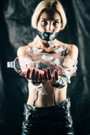 Plastic pollution. Defocused art portrait. Water shortage. Recyclable consumption. Environmental problems. Woman taped mouth body holding empty wrinkled bottle isolated dark blur background.の写真素材