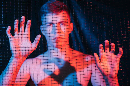 Man slavery. Defocused portrait. Human trafficking. Freedom protection. Disturbed scared shirtless male captive trapped behind cell in red blue neon light on blur dark background out of focus.の写真素材