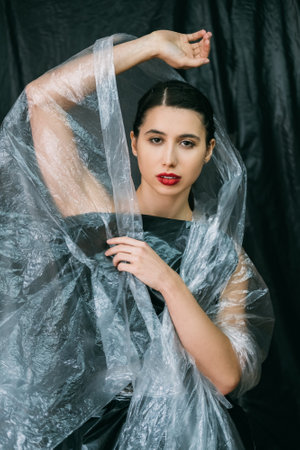 Beauty care. Plastic surgery. Anti aging procedure. Confident woman with perfect face skin night makeup red lips posing in wrinkled transparent polyethylene film veil isolated on dark background.の写真素材