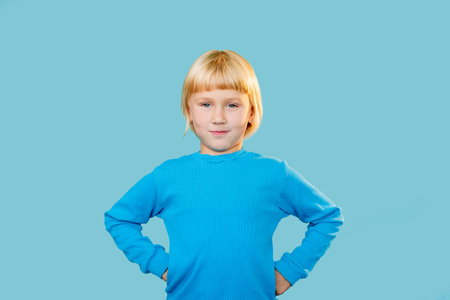 Naughty kid. Fun childhood. Positive lifestyle. Portrait of confident curious little girl standing in akimbo position looking at camera with sarcastic smile isolated on blue copy space background.の写真素材