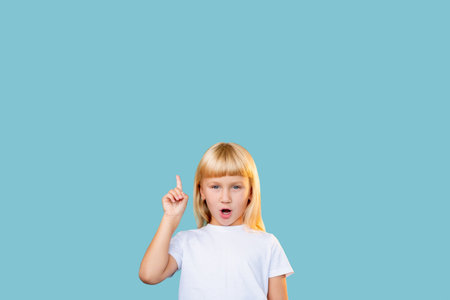 Solution idea. Advertising background. Aha moment. Knowledge discovery. Portrait of inspired surprised blonde little girl pointing up at invisible product information isolated on blue empty space.の写真素材