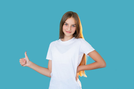 Satisfied girl. Like gesture. Well done. Approval sign. Portrait of happy enthusiastic cheerful young lady in white t-shirt showing thumb up smiling isolated on blue copy space background.の写真素材