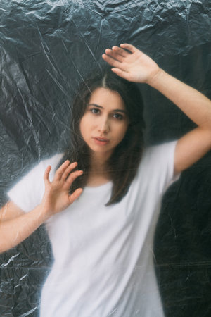 Female loneliness. Defocused portrait. Misery tragedy. Regret sorrow. Desperate sad woman trapped behind wrinkled texture transparent polyethylene film isolated on dark background out of focus.の写真素材