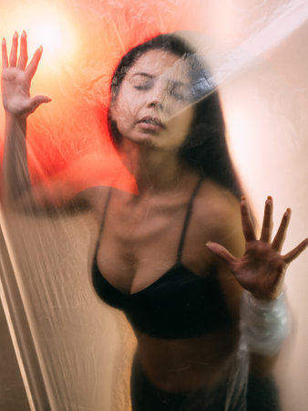 Female protection. Defocused portrait. Social isolation. Disturbed sensual woman in black lingerie with closed eyes trapped behind clear polyethylene film on orange sunset background out of focus.の写真素材