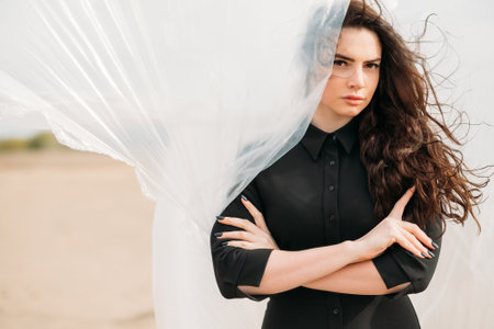 Fashion portrait. Feminine beauty. Success motivation. Confident brunette woman in black shirt with flying away long wavy hair standing with folded arms on windy sandy beach with white drape curtain.の写真素材