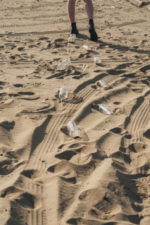 Plastic recycling. Waste reduction. Sustainable Earth. Ecology problem. Back view of unrecognizable woman legs walking away leaving used empty bottles trash abstract footprints on desert sand.の写真素材