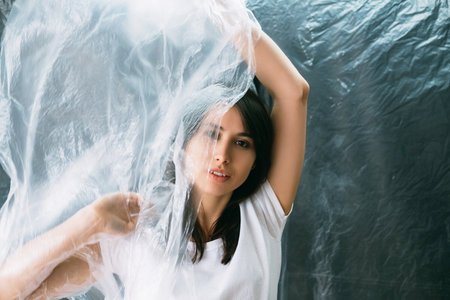 Beauty portrait. Aesthetic cosmetology. Anti aging procedure. Brunette woman with perfect skin covering face with wrinkled texture transparent polyethylene film isolated on dark copy space background.の写真素材