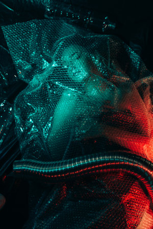 Environmental pollution. Defocused portrait. Save nature. Ecology problem. Hopeless woman trapped in waste dump under plastic bubble wrap exhaust pipe in night blue red neon light out of focus.の写真素材