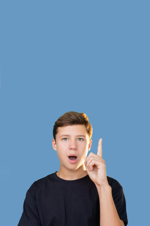 Creative idea. Advertising background. Aha moment. Insight discovery. Portrait of inspired surprised teen boy in black t-shirt pointing finger up looking at camera isolated on blue copy space.の写真素材