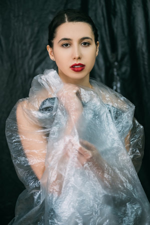 Beauty enhancement. Face contouring. Plastic surgery. Confident woman with perfect skin night makeup red full lips posing in wrinkled transparent polyethylene film isolated on dark background.の写真素材