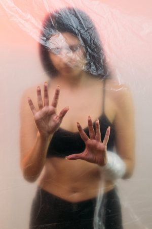 Pandemic self isolation. Defocused portrait. Lockdown loneliness. Depressed woman in black lingerie standing alone behind transparent polyethylene film on pastel pink background out of focus.の写真素材