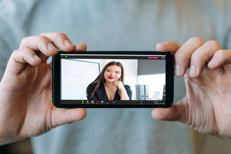 Video chat. Virtual meeting. Online communication. Digital office. Male hands showing successful business woman at workplace on mobile phone screen on blur background.の写真素材