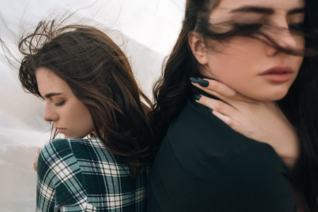 Gay love. Complicated relationship. Lgbt pride. Peaceful sensual lesbian women couple hiding feelings standing back to back with closed eyes in cold wind on light abstract cloudy sky background.の写真素材
