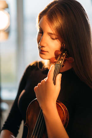 Female violinist portrait. Musical inspiration. Tranquility mind. Enjoying silence. Elegant smiling woman tenderly holding violin in hands warm lights.の写真素材