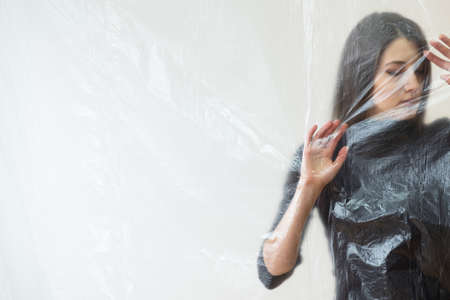 Sad woman. Defocused art portrait. Depression crisis. Social isolation. Psychology barrier. Helpless lady touching transparent plastic film isolated neutral copy space.の写真素材