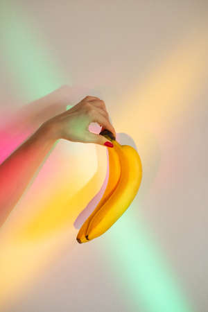 Two fresh bananas. Tropical food. Organic nutrition. Healthy diet. Unrecognizable woman holding yellow fruit bunch on blur rainbow overlay glowing neon lights background.の写真素材