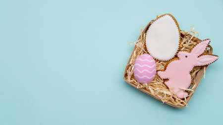 Easter gift. Holiday food ornament. Homemade pastry decoration. Sweet pink white gingerbread bunny biscuit painted egg set for girl in rustic basket with straw isolated on blue copy space background.の写真素材