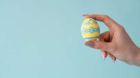 Easter egg. Festive ornament. Handmade food decoration. Female hand holding yellow color traditional spring holiday symbol with folklore pattern isolated on blue copy space background.の写真素材