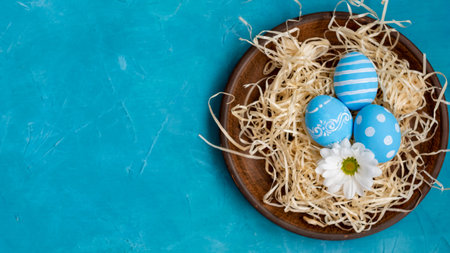 Easter banner. Holiday food decoration. Festive composition. Painted eggs with creative modern pattern Spring white flower in rustic plate with straw on blue textured copy space background.の写真素材