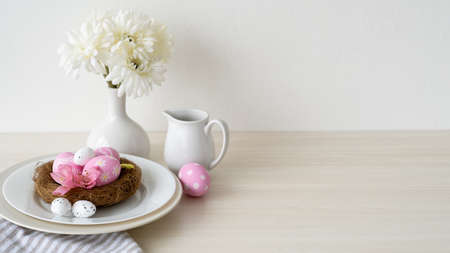 Easter dinner. Festive table. Holiday celebration. Banquet service. Pink color painted eggs in nest served in plate with white Spring flower bouquet arrangement on light copy space background.の写真素材