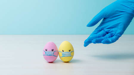 composition. Medical care. Risk prevention. Doctor hand in blue protective glove offering help to painted Easter egg couple in face mask on light conceptual empty space background.の写真素材