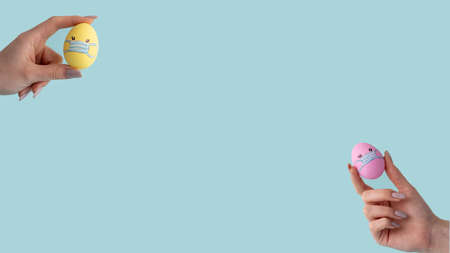 Pandemic banner. Coronavirus quarantine. Lockdown self isolation. Set of 2 yellow pink color Easter egg with face in mask design in female hand isolated on blue empty space conceptual background.の写真素材