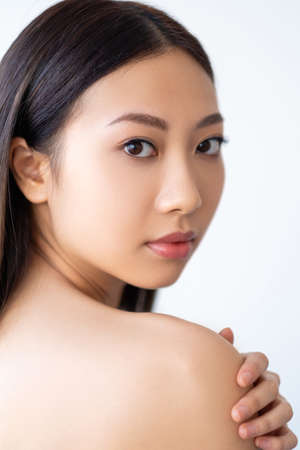 Beauty care. Skin treatment. Spa therapy. Wellness freshness. Portrait of sensual Asian woman with perfect smooth face nude makeup touching bare shoulder isolated on white background.の写真素材