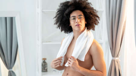 Male body care. Morning hygiene. Metrosexual lifestyle. Handsome confident shirtless curly guy in eyeglasses with smooth face skin white towel after shower in light modern bathroom.の写真素材
