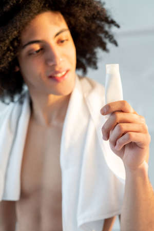 Man skincare. Cosmetic product advertising. Facial treatment. Cheerful handsome shirtless guy brown curly hair towel after shower holding white mockup cream tube in hand on light defocused background.の写真素材