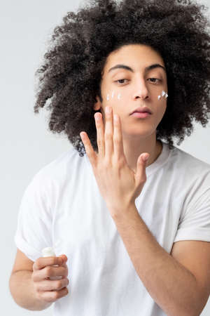 Male skincare. Face moisturizing. Cosmetic product. Confident handsome guy with long brown curly hair in white t-shirt applying facial cream on smooth skin isolated on light background.の写真素材