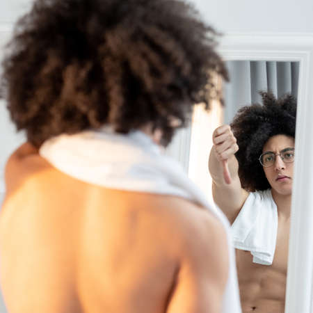 Unhappy man. Body shaming. Low self-esteem. Dissatisfied handsome shirtless guy in eyeglasses long brown curly hair towel on bare shoulders showing thumb down gesture to reflection in mirror.の写真素材
