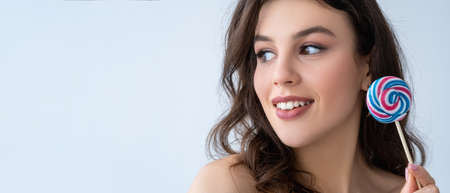 Beauty banner. Skin care. Aesthetic cosmetology. Cheerful curious woman with flawless face white toothy smile colorful lollipop looking at copy space isolated on light advertising background.の写真素材