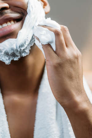 Male skincare. Facial hygiene. Morning grooming. Cheerful African man applying white shaving foam on dark face skin with hand smiling isolated on light defocused background.の写真素材