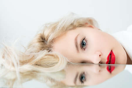 Vision correction. Eye laser surgery. Eyesight treatment. Ophthalmic procedure. Art portrait of blonde woman face with red lips makeup with blur mirror reflection isolated on light background.の写真素材