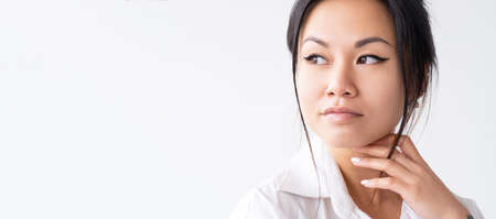 Beauty portrait. Skincare banner. Aesthetic cosmetology. Pensive Asian woman with perfect face skin nude makeup isolated on white empty space commercial background.の写真素材