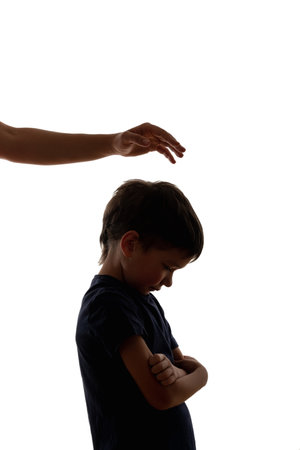 Offended kid. Family conflict. Domestic violence. Punishment abuse. Sad little boy crossing hands on chest with unrecognizable parents hand under head isolated on white.の写真素材