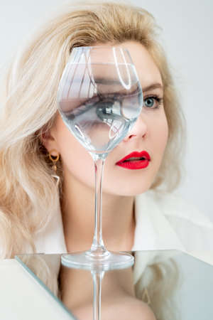 Vision correction. Eye surgery. Eyesight laser therapy. Optical illusion. Art portrait of blonde woman face with red lips looking through blur water glass prism magnifier isolated on light background.の写真素材