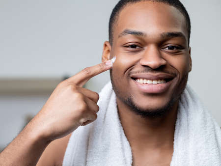Male skincare. Facial treatment. Cosmetic product advertising. Confident cheerful African man with white towel showing pointing with finger cream on dark face skin smiling on light background.の写真素材