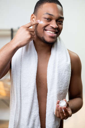 Man skincare. Cosmetic product. Anti aging facial treatment. Happy satisfied shirtless African guy with towel applying face cream on dark skin with finger from small jar after shower smiling.の写真素材