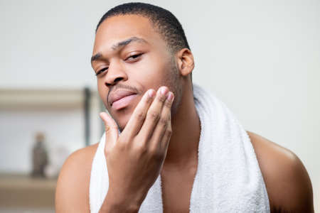 Male grooming. Skin care. Facial treatment. Beard moisturizing. Confident shirtless African man with white towel touching face with hand after cleansing.の写真素材