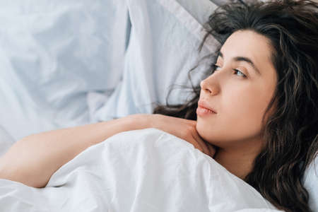 Morning melancholy. Weekend loneliness. Home depression. Pensive tired sad brunette woman lying in bed alone with white blanket.の写真素材