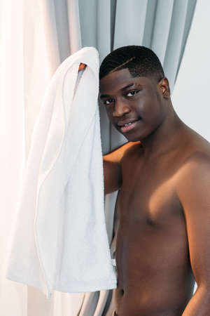 Shirtless black man. Hygiene treatment. Bathroom routine. Male body care. Smiling athletic african guy wiping hair white towel looking at camera light home interior.の写真素材