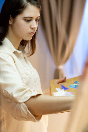 Female painter. Art studio. Drawing lesson. Creative hobby. Inspiration muse. Calm woman painting on drawing easel with paints palette in light classroom.の写真素材