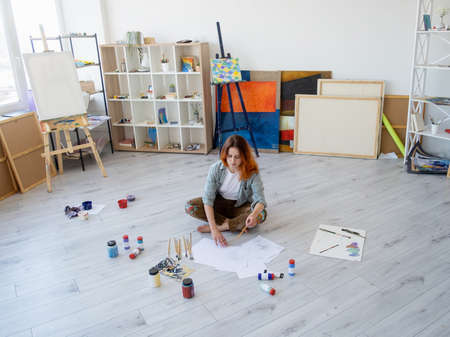 Painting art. Artist lifestyle. Creative hobby. Inspired relaxed woman painter drawing sketches with pencil sitting on floor in modern light studio with paintbrushes supplies easel artwork collection.の写真素材