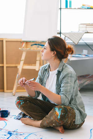 Painting art. Creative lifestyle. Talent imagination. Inspired pensive female artist sitting with paintbrush cross-legged barefoot on colorful abstract artwork on floor in light workshop.の写真素材