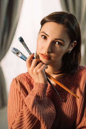 Artist portrait. Art inspiration. Creative lifestyle. Pensive curious female painter with brushes in cozy workshop on light window background.の写真素材